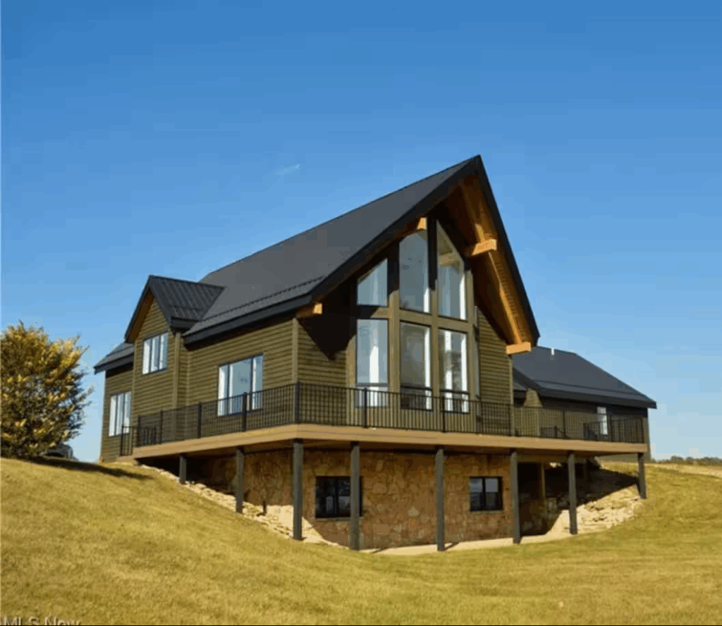 Exterior view of White Pines Lodge, a large modern vacation home in Coshocton County, Ohio, featuring floor-to-ceiling windows, a wraparound deck, and scenic hilltop surroundings under a clear blue sky.